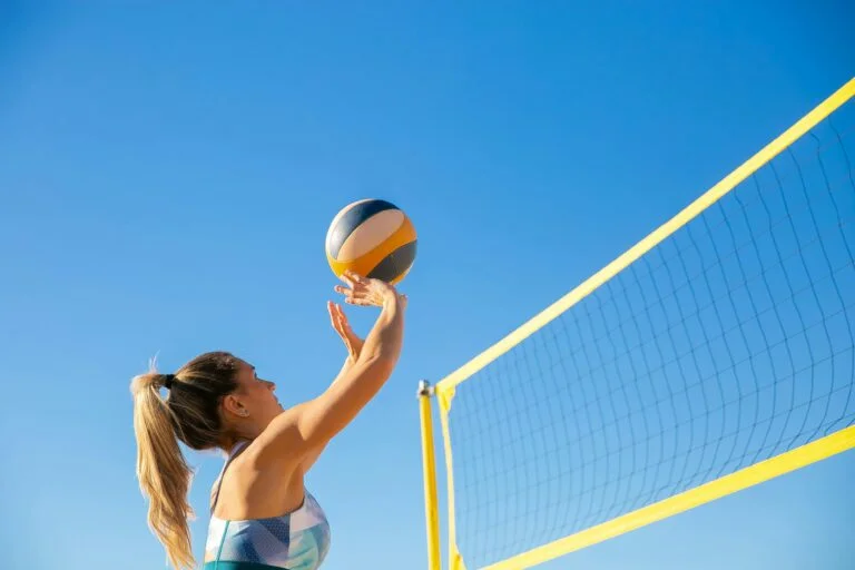 A woman plays volleyball on a sunny day, reaching to hit the ball over the net.