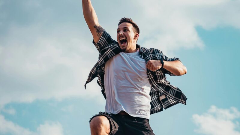 A joyful young man jumps midair with clouds and blue sky in the background, exuding energy and freedom.