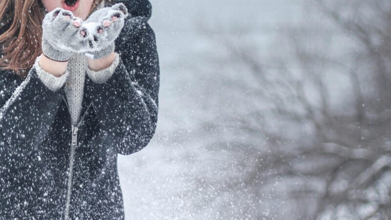 Woman in cozy winter clothing blowing snowflakes with excitement outdoors in a snowy setting.