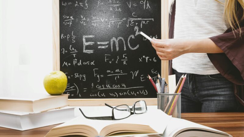 Woman writing physics equations on a blackboard with books and an apple on the desk.