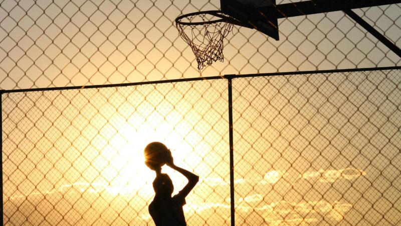 Silhouetted athlete playing basketball on an outdoor court with a vibrant sunset backdrop.